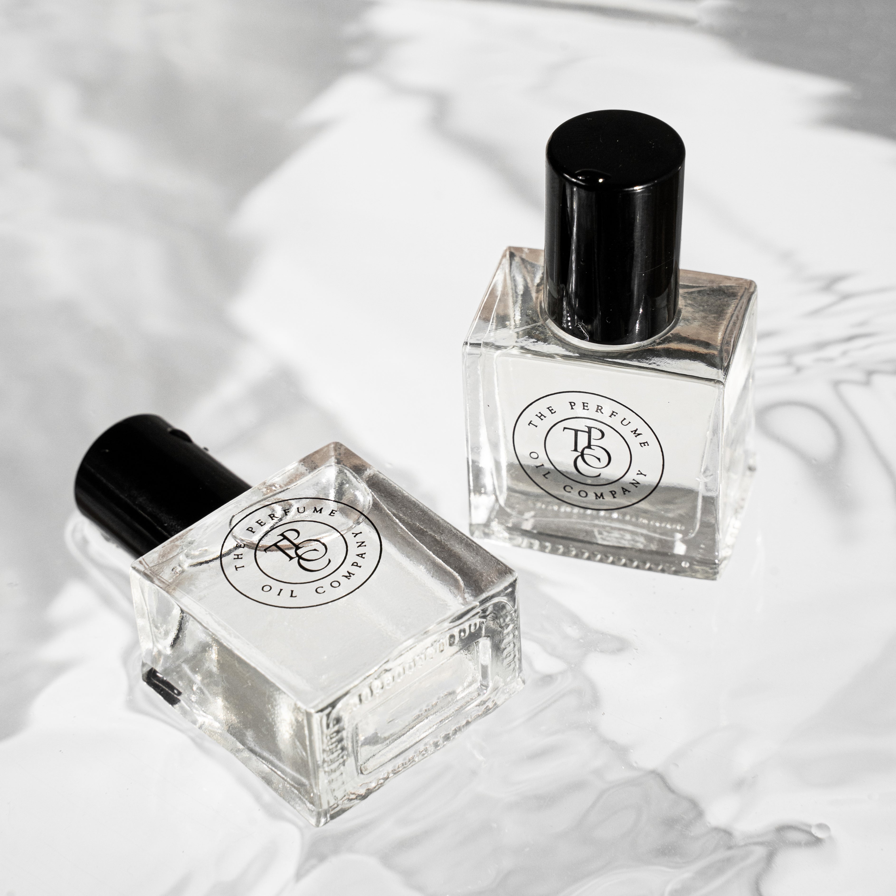Two minimalist glass bottles of The Perfume Oil Company fragrance, featuring sleek black caps and circular logo labels, resting on a softly rippled, light-reflective surface.